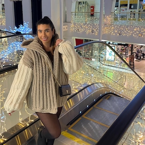 Könnte beinhalten: Eine Frau trägt einen beige-weißen Strickcardigan mit einem schwarzen Minirock und schwarzen Strumpfhosen. Sie steht auf einer Rolltreppe in einem Einkaufszentrum, das mit Weihnachtslichtern geschmückt ist.