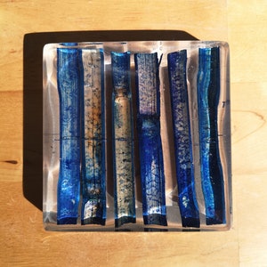 May include: A square, clear glass tray with a blue and white design. The design features five vertical stripes of blue and white, resembling wood grain.