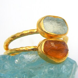 May include: Two gold rings with a hammered texture. The top ring features a light blue gemstone, while the bottom ring has an orange gemstone.