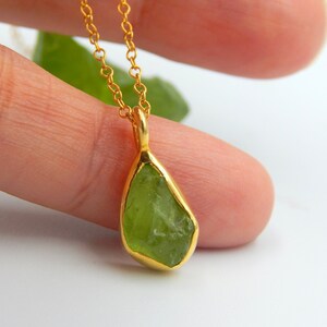 May include: A gold chain necklace with a green peridot pendant. The peridot is a raw, uncut stone with a natural, irregular shape.