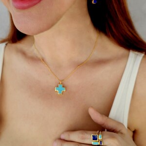 May include: A gold necklace with a turquoise cross pendant. The necklace is worn by a person with long brown hair. The person is also wearing a gold ring with turquoise and blue stones.
