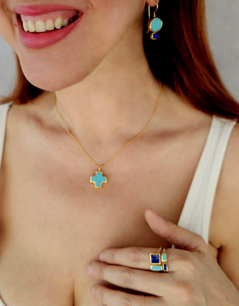 May include: A gold necklace with a turquoise cross pendant. The necklace is worn by a person with long brown hair. The person is also wearing a gold ring with turquoise and blue stones.
