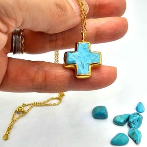 May include: A gold chain necklace with a turquoise cross pendant. The cross is made of gold and has a turquoise stone in the center. There are several turquoise stones scattered on a white surface.