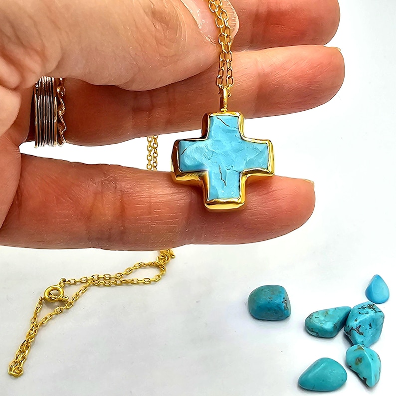 May include: A gold chain necklace with a turquoise cross pendant. The cross is made of gold and has a turquoise stone in the center. There are several turquoise stones scattered on a white surface.