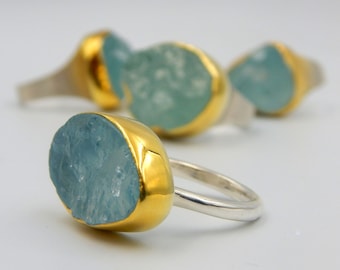 Puede incluir: Un anillo con una piedra preciosa de aguamarina azul claro engastada en una banda dorada. El anillo está sobre un fondo blanco.