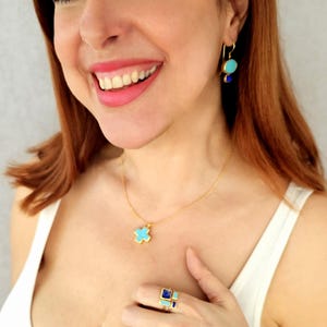 May include: A woman wearing a white tank top and gold jewelry with turquoise and blue stones. The jewelry includes a necklace with a turquoise cross pendant, a ring with turquoise and blue stones, and earrings with turquoise and blue stones.