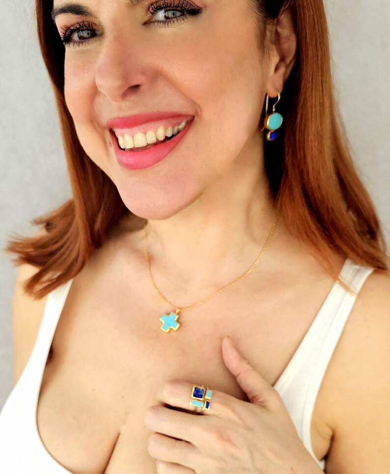 May include: A woman wearing a white tank top and gold jewelry with turquoise and blue stones. The jewelry includes a necklace with a turquoise cross pendant, a ring with turquoise and blue stones, and earrings with turquoise and blue stones.