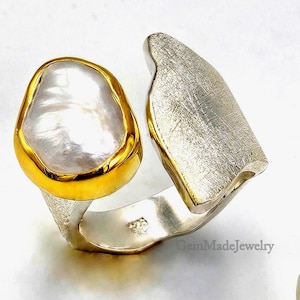 Op de afbeelding: Een unieke ring met een grote, onregelmatige parel gezet in een gouden zetting. De ringband is zilverkleurig met een getextureerde, geborstelde afwerking aan de zijkant. Het ontwerp is modern en opvallend.