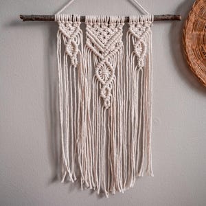 Op de afbeelding: Een crèmekleurige macramé wandhanger met een geometrisch ontwerp, opgehangen aan een natuurlijke houten tak. De ingewikkelde knoopwerk creëert een textuurpatroon, met lange franjes aan de onderkant. Een gevlochten mand is zichtbaar op de achtergrond.