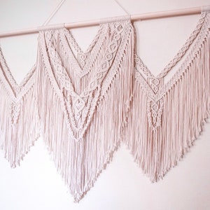 以下が含まれることがあります： 白とクリーム色のマクラメ壁掛け3点セット。壁掛けはコットンコードで作られており、フリンジのデザインが施されています。木製ダウエルロッドに吊り下げられています。