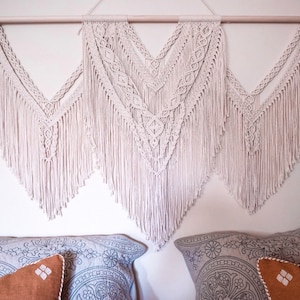 May include: A white macrame wall hanging with a fringe border. The hanging is made up of three sections, each with a different pattern. The hanging is mounted on a wooden dowel rod.
