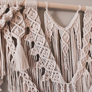 K&ouml;nnte beinhalten: Ein beigefarbener Makramee-Wandbehang mit einem Holzstab oben. Der Wandbehang besteht aus verschiedenen Knoten und Mustern, darunter eine Quaste.