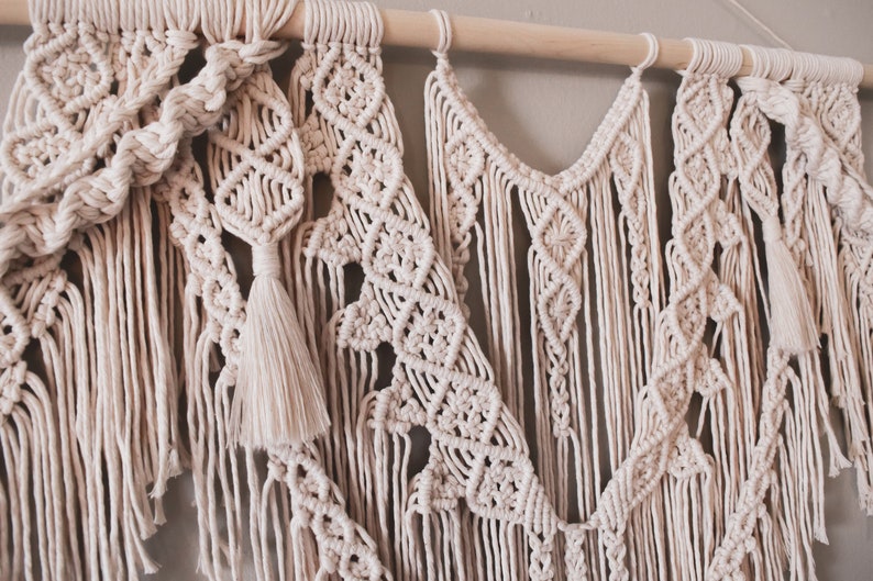 K&ouml;nnte beinhalten: Ein beigefarbener Makramee-Wandbehang mit einem Holzstab oben. Der Wandbehang besteht aus verschiedenen Knoten und Mustern, darunter eine Quaste.