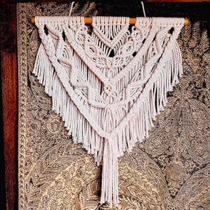 May include: A white macrame wall hanging with a triangular shape and a fringe border. The hanging is made of cotton cord and has a wooden dowel at the top.