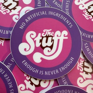 Puede incluir: Una colección de pegatinas redondas con borde morado y centro rosa. Las pegatinas presentan el texto "The Stuff" en una fuente blanca estilizada, y las frases "No Artificial Ingredients" y "Enough is Never Enough."