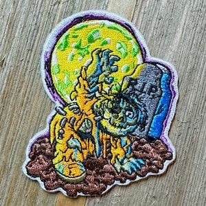 May include: A yellow and blue embroidered patch of a zombie rising from a grave with a full moon in the background. The zombie has a green face and is holding a tombstone that says "RIP".