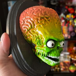 May include: A wall-mounted plaque featuring a three-dimensional alien head sculpture with a large, textured, brain-like cranium that transitions from orange to green. The figure has prominent white eyes with black pupils and a green jaw with white teeth.