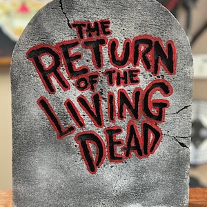 May include: A gray tombstone with a crack down the middle. The words "THE RETURN OF THE LIVING DEAD" are painted in black and red letters.
