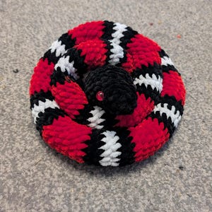 Peut inclure: Un serpent en peluche enroulé, fabriqué en fil doux, présentant un motif répétitif de bandes rouge vif, noires et blanches. Le serpent possède une tête noire unie avec un seul œil rouge brillant visible, disposé en position circulaire.