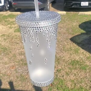 May include: A clear, frosted tumbler with a bedazzled lid and base. The lid and base are covered in small, sparkling rhinestones. Rhinestones also drip down the side of the cup. A clear straw is inserted into the cup.
