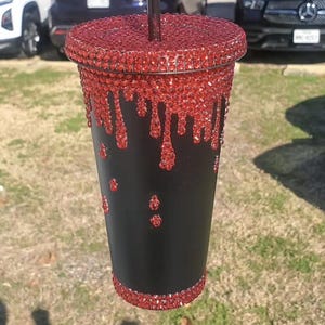 May include: Black tumbler with a red rhinestone lid and dripping design. The red rhinestones are also on the bottom rim and in a few spots on the cup. A black straw is inserted into the lid. The cup is held up in front of a blurred background.