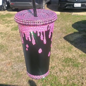 May include: A black tumbler cup adorned with pink rhinestones. The lid and base are fully covered in rhinestones, while the cup features a dripping rhinestone design. A black straw is inserted into the lid. The cup is held outdoors in front of a blurred background.
