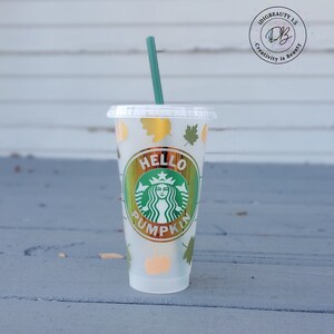 Puede incluir: Un vaso reutilizable de plástico transparente con un logotipo de Starbucks dorado y verde y el texto "Hello Pumpkin". El vaso tiene una pajita verde y está decorado con hojas de otoño doradas y verdes.