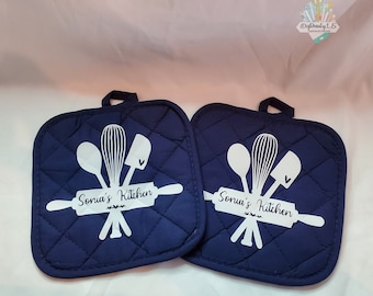 May include: Two navy blue quilted pot holders with white text and a design of kitchen utensils, including a rolling pin, a whisk, a spatula, and a spoon. The text on each pot holder reads "Sonia's Kitchen".