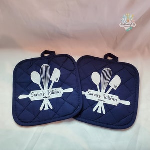 May include: Two navy blue quilted pot holders with white text and a design of kitchen utensils, including a rolling pin, a whisk, a spatula, and a spoon. The text on each pot holder reads "Sonia's Kitchen".