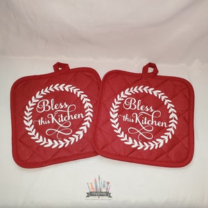 May include: Two red quilted pot holders with white trim and the text "Bless this Kitchen" in a white wreath design.