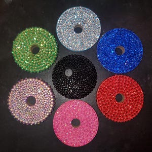 May include: A collection of seven bedazzled, circular bottle stoppers. Each stopper is a different color, including green, silver, blue, black, red, pink, and iridescent. The stoppers have a hole in the center.