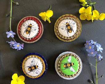 May include: Four round bottle cap magnets with a bee design. The magnets are red, white, blue, and green. The bees are yellow and black.