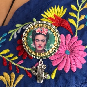 May include: A Frida Kahlo brooch with a silver cactus charm. The brooch features a portrait of Frida Kahlo with a floral crown, surrounded by a metallic border. The background is a blue embroidered fabric.