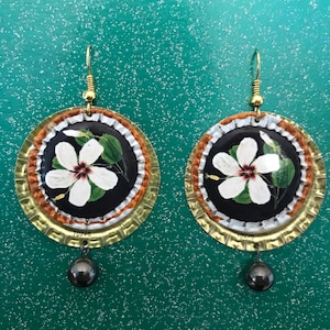 May include: A pair of handmade earrings featuring a white hibiscus flower design on a black background. The earrings are crafted from repurposed bottle caps with gold-colored hooks and a dark gray bead. The number "10276" is visible on the bottom of each earring.