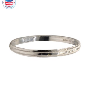 May include: A silver-toned bangle bracelet featuring a polished finish with a central textured band. The interior of the bracelet is engraved with the text 'MADE IN USA'. The design is circular and metallic, showcasing a clean, reflective surface.
