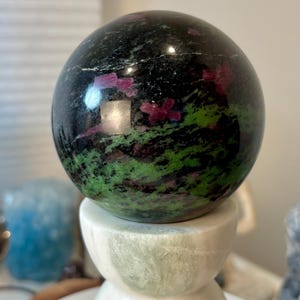 May include: A polished spherical stone featuring a dark black base with vibrant green mineral inclusions and scattered magenta-colored crystalline patches, resting atop a smooth, cylindrical white and pale green marble pedestal stand.