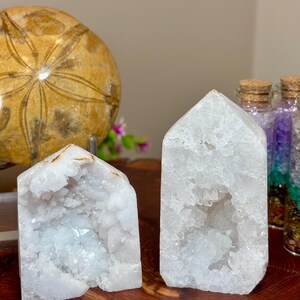 May include: Two white quartz crystal points with hollowed, crystalline interiors. The crystals feature jagged, translucent surfaces and pointed tops. To the left is a round, tan, textured object, and to the right are glass vials filled with colorful stones.
