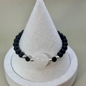 May include: A beaded bracelet featuring black and white gemstones. The bracelet has a central large white stone, flanked by smaller black stones and silver-colored beads. The bracelet is displayed on a white, cone-shaped stand.