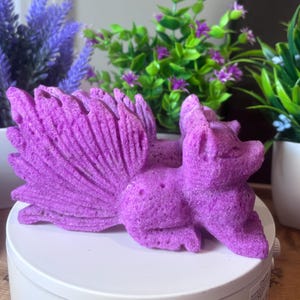 May include: A vibrant purple fox sculpture with a multi-layered tail, resting on a white surface. The fox is detailed with textured fur and is surrounded by potted plants with purple and green foliage.