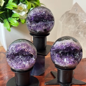 May include: Three spherical amethyst geodes with deep purple crystalline interiors, each mounted on a black metal stand. Two stands have circular bases, while one features a star-shaped base. The stones display natural jagged crystal formations.