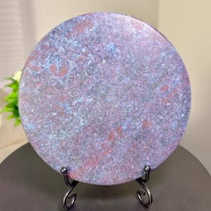 May include: A circular stone slab featuring a mottled pattern of deep blue, purple, and reddish-brown mineral flecks. The polished surface has a smooth, reflective texture and is displayed upright on a black metal wire stand with curved feet.