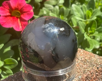 May include: A polished, black sphere sits on a clear, round base, reflecting the sky and surrounding greenery. The sphere is placed on a weathered, reddish-brown brick surface, with vibrant pink flowers and green foliage in the background.