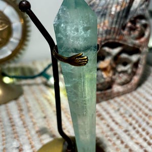 May include: A tall, light green fluorite crystal is held by a bronze-colored metal stand. The stand features a round base and a curved arm with a hand-shaped detail that embraces the crystal. The crystal is approximately 6 inches tall.