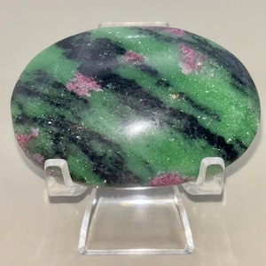 May include: Oval-shaped zoisite stone with ruby inclusions. The stone features a vibrant green base with black veining and small patches of pink ruby crystals. The polished surface reflects light, highlighting the natural patterns and colors. The stone is displayed on a clear acrylic stand.
