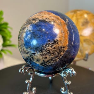 May include: A polished spherical stone featuring deep blue, tan, and black mineral inclusions. The sphere rests on a silver-toned metal tripod stand with ornate, curved legs that provide a stable base for the rounded mineral specimen.