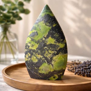 May include: A teardrop-shaped stone sculpture with a polished surface. The stone features a striking pattern of green and black hues. The sculpture is displayed on a wooden tray, showcasing its natural beauty. A decorative piece for home decor.