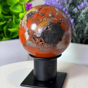 May include: A polished, spherical gemstone with a vibrant mix of red, black, and gold hues. The orb is mounted on a simple black metal stand with a square base, showcasing the natural patterns and textures of the stone. The gemstone is a decorative item.