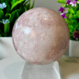 May include: A large, spherical, rose quartz crystal ball. The sphere is a soft pink color with subtle white veining. It rests on a clear, square selenite crystal base. Green and purple plants are in the background.