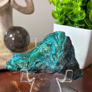 May include: A raw specimen of chrysocolla and malachite mineral resting on a clear acrylic display stand. The stone features vibrant bands of turquoise and deep green, contrasted by a rough, dark, and textured rocky exterior surface.