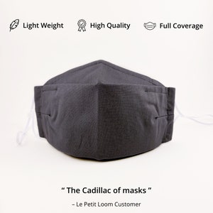 May include: A dark gray reusable face mask with white elastic ear loops. The mask has a pleated design and is made of a soft, breathable fabric. The text "The Cadillac of masks" and "Le Petit Loom Customer" is printed below the mask.